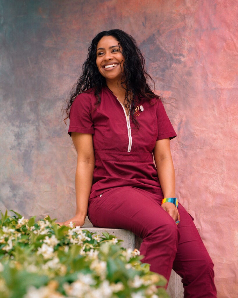 Black woman in scrubs, a nurse at the Center, posing outdoors in front of a rainbow backdrop.