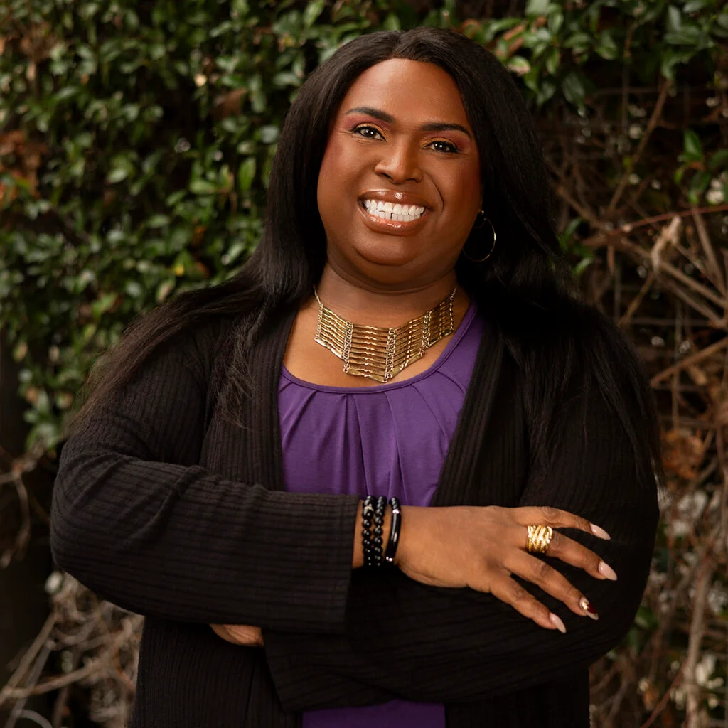 Trans Wellness Center staff member smiles at the camera, standing in front of green foliage.
