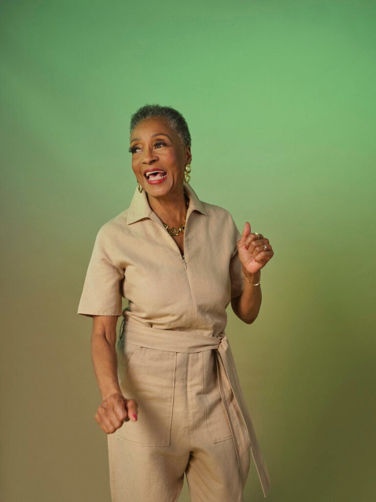 A Black senior woman dances; she is wearing a khaki jumpsuit and there is a green backdrop behind her.