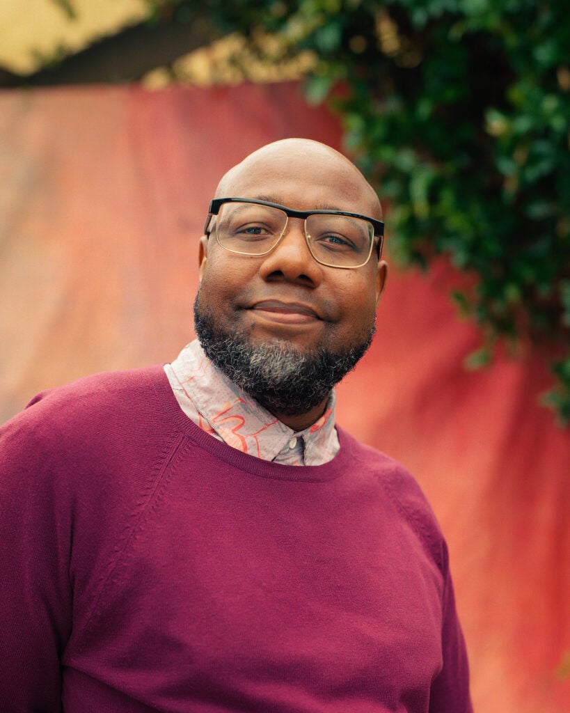 An elder, Black trans man poses in front of a salmon-covered backdrop outdoors. He wears glasses, a magenta sweater, and has light facial hair.
