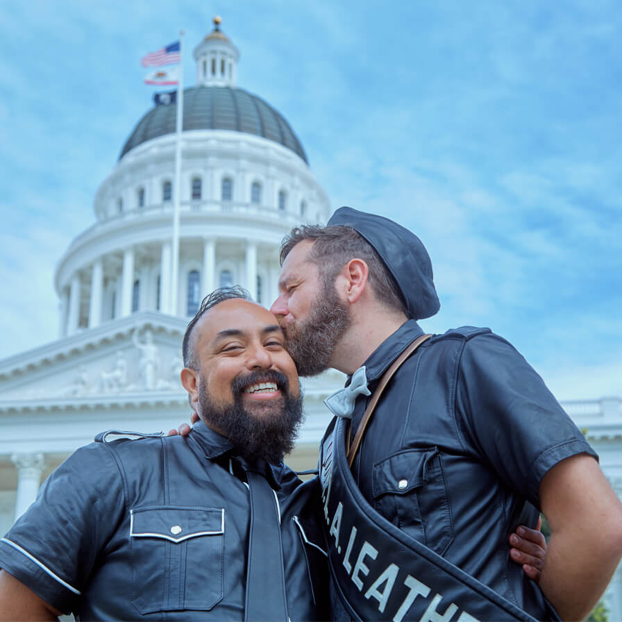 Two men wearing leather outside of California state capital, one man gives other a kiss on head