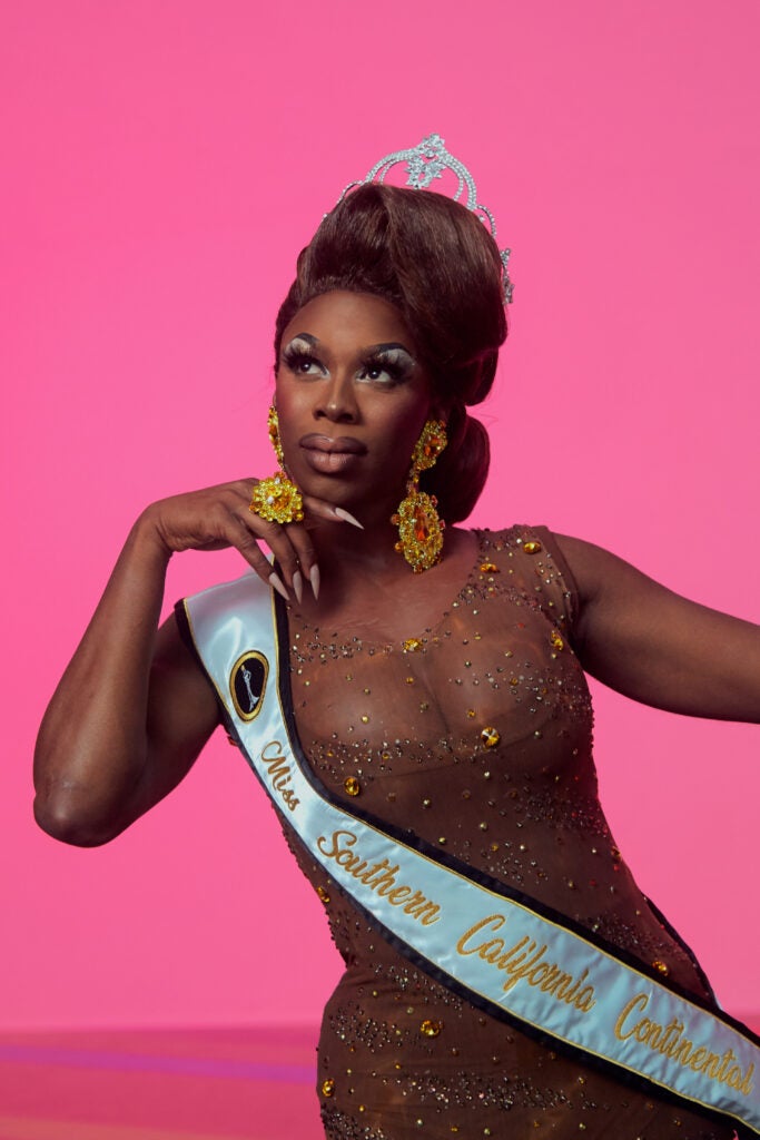 Drag queen Honey Davenport pose against a bright pink background—she is wearing a tiara, a sash, and a crystalline dress. Her hair is styled in an updo.