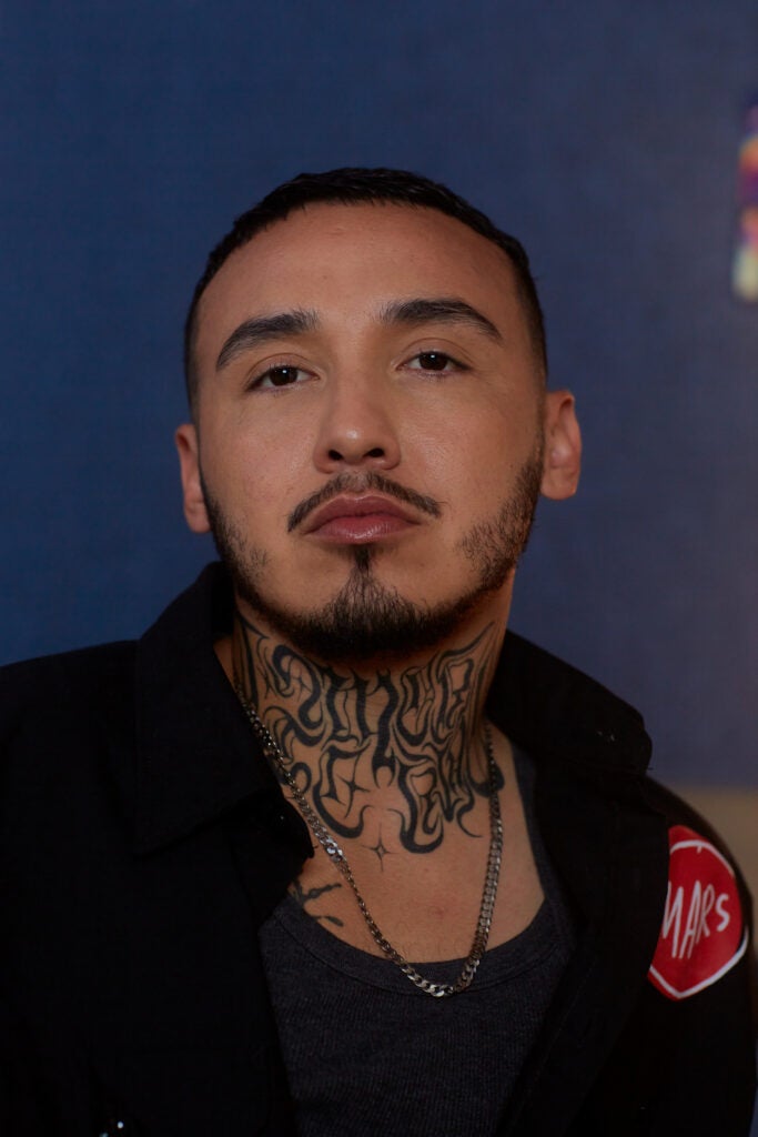 A Latinx trans man with neck tattoos and facial hair looks directly at the camera against a dark blue background