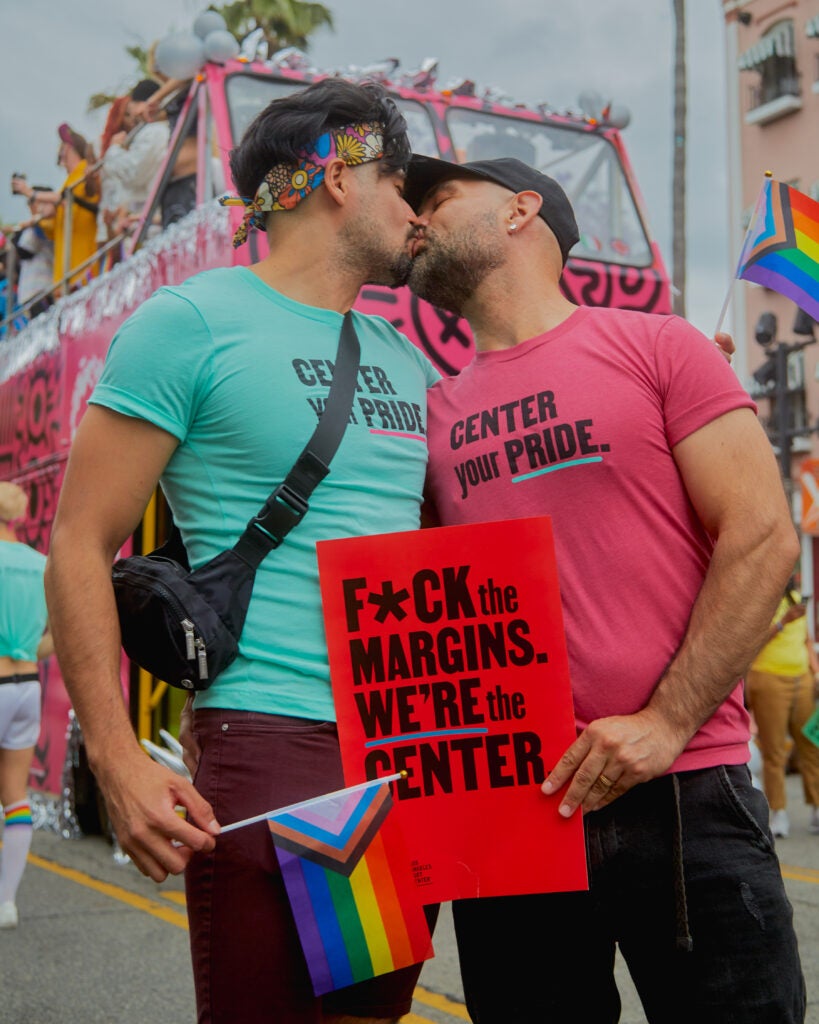 A gay Latino couple kisses at a Pride parade, wearing bright colored t-shirts that say "Center Your Pride" and holding a sign that says, "Fuck the Margins, We