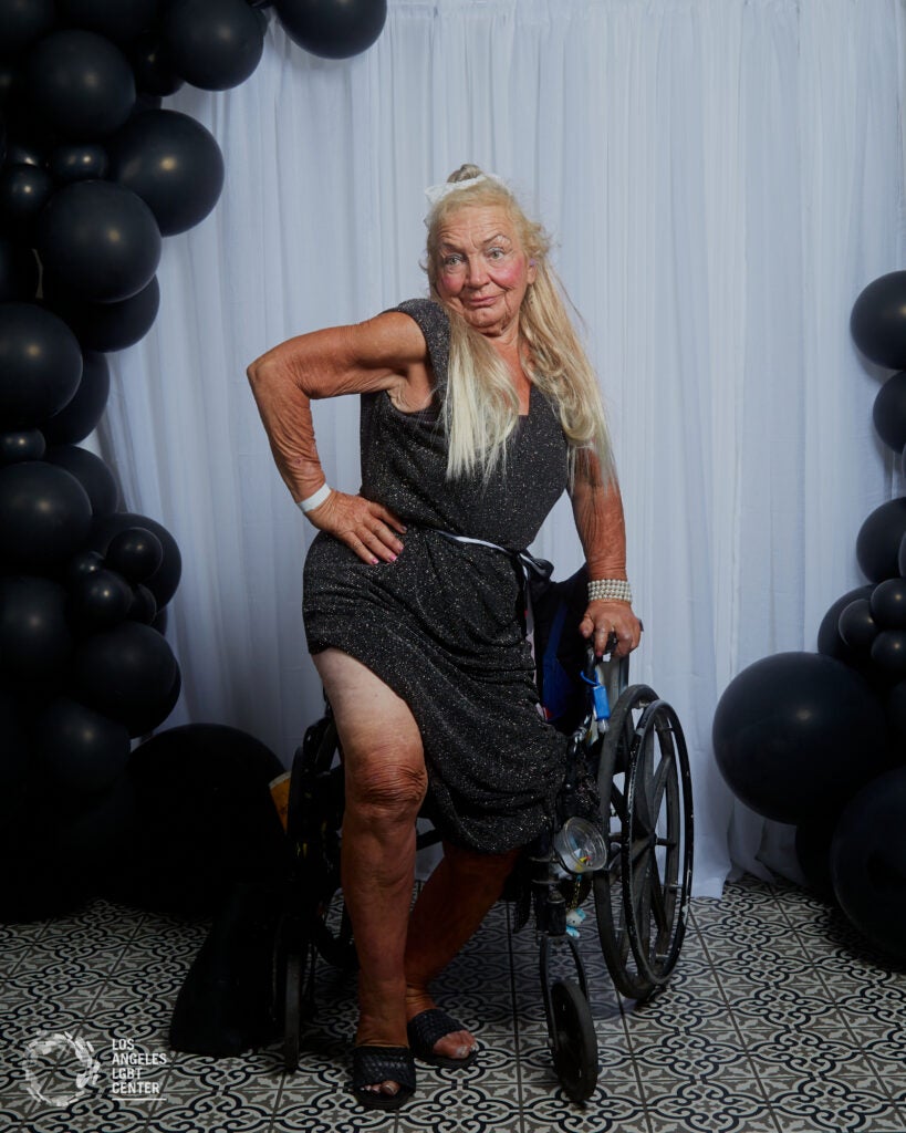 Woman with long hair and black, semi-formal dress poses with her leg prominently displayed. She uses a wheelchair.