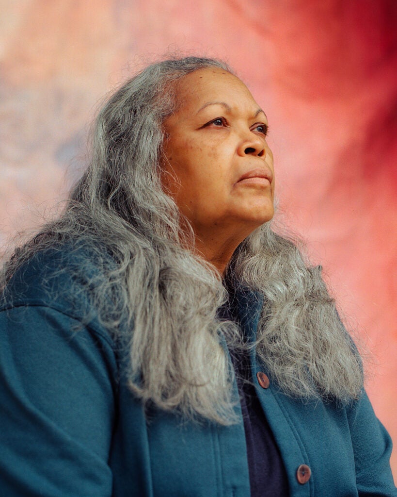 A Black LGBTQ+ senior woman looks away from the camera; there is a rainbow backdrop behind her. She has long, gray hair.