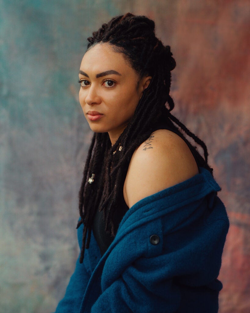 A Black woman poses against a rainbow, watercolor backdrop. She wears long twists, and a blue sweater that is draped, revealing her shoulder.